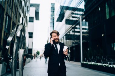 Şehir caddesinde yüksek binalar arasında duran ve kahve içerken akıllı telefondan konuşan zarif resmi kıyafetler giyen pozitif sakallı bir çalışan.