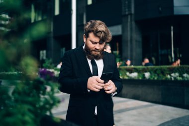 Resmi takım elbiseli ve kravatlı ciddi sakallı bir adam modern binanın yanında dikiliyor ve şehirde cep telefonuyla sohbet ediyor.