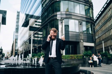 Resmi takım elbiseli ve kravatlı iyimser sakallı işadamı modern binanın yanında duruyor ve başarılı bir anlaşmayı kutlarken akıllı telefondan arıyor.