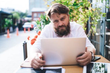 Ciddi sakallı, beyaz tişörtlü, tahta masada oturan ve dizüstü bilgisayar projesinde çalışırken kameraya bakan serbest bir erkek.