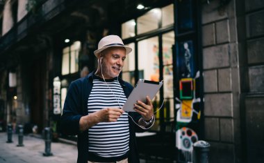 Elektronik kulaklıklı gülümseyen beyaz erkek turist dijital tablet ve 4g internet bağlantısı kullanarak uluslararası video araması yapıyor. 60 'lı yaşlardaki mutlu adam cihaz web sitesinde haber okuyor.