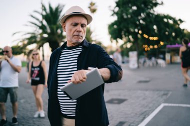 Elde dokunmatik pedlerle emekli olmuş modaya uygun giyinmiş erkek turist, sokak okumalarında elinde dijital tablet bulunan saman şapkalı yaşlı Kafkasyalı adam, giyilebilir akıllı gözetimi kontrol ediyor.