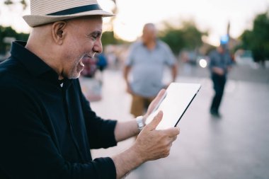 Hasır şapkalı yetişkin bir adam dokunmatik ekranda mesajları kontrol ediyor ve heyecanlı, mutlu, şok olmuş bir erkek turist ağı web sitesinde dijital cihazda 4g internet kullanarak izlenim haberi veriyor.