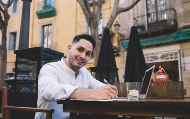 Beyaz gömlekli neşeli genç adam deftere yazı yazıyor ve sokak kafesinden dizüstü bilgisayarla kameraya bakıyor.