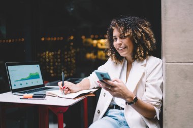 Bilgisayarında projeksiyon ve web diyagramlarıyla uzaktan çalışan mutlu kadın bilişim uzmanı elinde akıllı telefon cihazıyla kameraya gülümseyen ve serbest yaşam tarzının keyfini çıkaran neşeli kadın.