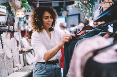 Moda mağazasında kıyafet seçen çekici modacı kadın alışveriş için karar vermeye odaklanmış, yetenekli tasarımcının galerisinde şık kıvırcık kadın kıyafetleri giyip moda modasına uygun bir görünüm yaratıyor.
