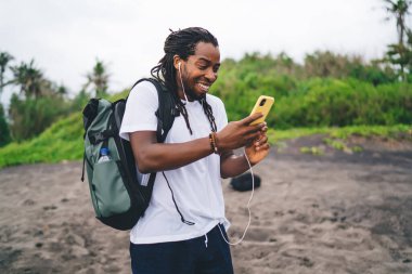 Tropikal yeşil ağaçların yanında kumda duran ve arka planda bulanık kulaklıklar kullanarak akıllı telefon aracılığıyla sohbet eden heyecanlı siyah erkek turist.