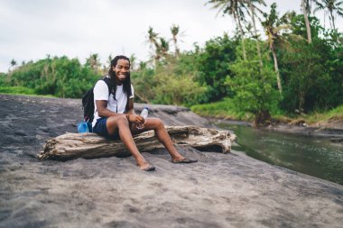 Uzun boylu, neşeli Afro-Amerikan erkek ağaç gövdesinde oturuyor ve nehrin kenarında gülümserken kameraya bakıyor.