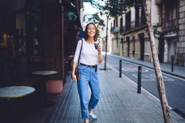 Güzel bir kasabada sokakta yürürken elinde defter ve kahveyle kameraya gülümseyen mutlu bir kadın.