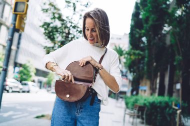 Orta yaşlı zarif bir kadın güneşli bir yaz gününde modern şehir caddesinde dikilirken kahverengi deri bir çanta açıyor.