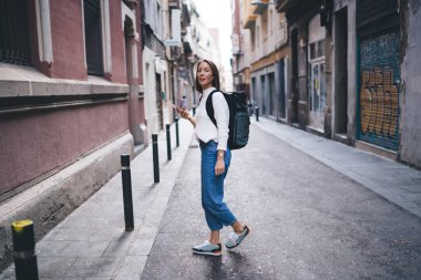 Sırt çantalı milenyum turisti, şehir merkezlerini ve caddeleri gezen cep telefonu teknolojisi olan çekici bir kadın, gezintiler sırasında konumu aramak için akıllı telefon uygulaması kullanıyor.