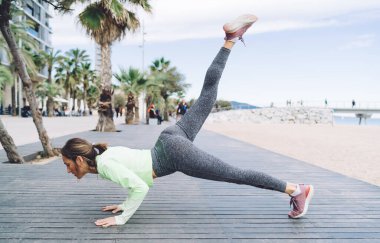 Tam boy orta yaşlı bayan sporcunun yan görünüşü. Yoga pozisyonu alıyor, dirsek eğiyor ve açık deniz havasında bacak kaldırıyor.