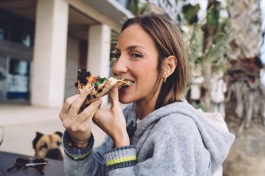Side view of middle aged female in warm sweater eating piece of pizza in street terrace of restaurant in city
