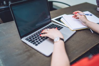 High angle of anonymous female in wristwatch sitting at wooden table and browsing laptop while taking notes in copybook during work