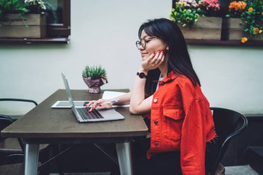 Side view of content Asian female freelancer in casual wear sitting at wooden table and working on laptop on terrace