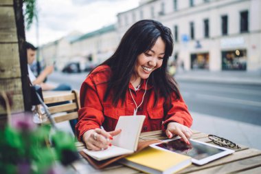 Positive young Asian brunette in trendy denim jacket surfing net on tablet and writing down content plan for blog while sitting at small table in outdoor cafe