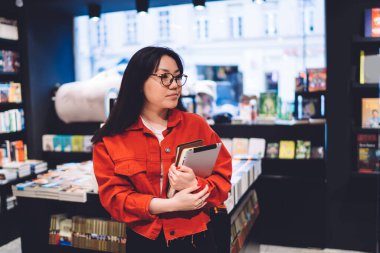 Unrecognizable female worker with tablet and notebook in hands spending time alone in bookstore while search for new amusing reading