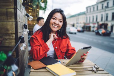 Young cheerful Asian lady in trendy denim jacket surfing net on tablet and smiling candidly at camera while enjoying free time in street cafe