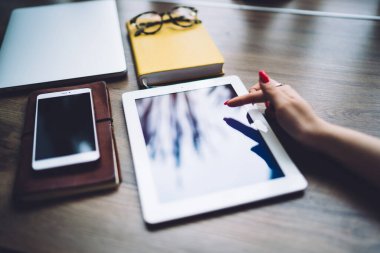 Anonymous female employee reading important information on screen and sitting at desk with planners while working on new business project