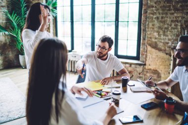 Multiracial male and female colleagues discussing business ideas enjoying collaborative meeting in coworking space, young hipster guys talking while learning togetherness using paper reports indoors
