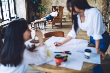 Blurred content females in white shirts working on poster on round table near concentrated male friend sitting with legs crossed on cozy leather chair in spacious loft studio