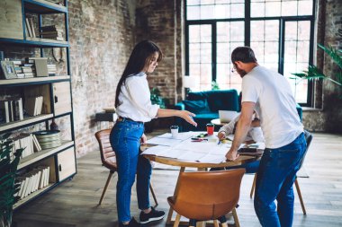 Young community members wearing similar clothes gathering at table with papers and markers while creating new design project in contemporary interior