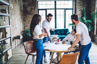 Group of creative young multiracial entrepreneurs in casual clothes sharing ideas while working together around table with documents in modern office