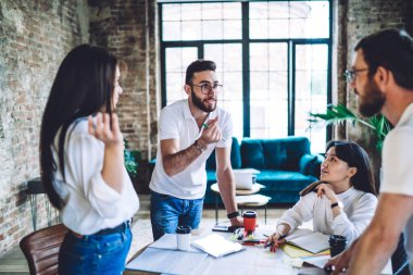 Serious young ethnic male employee in casual outfit and eyeglasses disputing  with colleague while working together on project in contemporary office