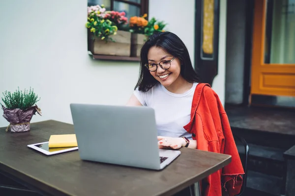 身穿休闲装和眼镜的快乐的亚洲女性自由职业者坐在室外阳台的木制桌旁 一边微笑一边浏览笔记本电脑 — 图库照片