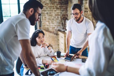 High angle of concentrated young multiethnic business partners in casual outfits cooperating on project while gathering at round table in creative workspace