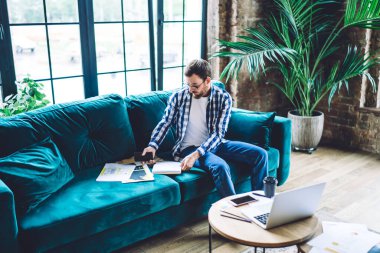Full length of bearded guy in glasses reading notes in notebook while sitting on comfortable sofa near panoramic window in bright room