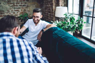 High angle of happy bearded male colleagues in casual outfits and glasses smiling and talking about business project in office