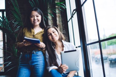 Low angle of positive multiracial female in casual clothes smiling and looking at camera while sitting near big window and browsing modern tablet