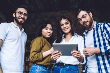 Low angle of positive multiracial coworkers in casual clothes standing close to each other and using modern tablet while working on interesting startup project together