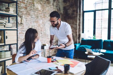 Cheerful multiethnic partners working on project in modern office and discussing details while searching for information from book during harmonious work