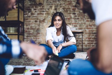 Casual woman smiling and sitting in lotus pose on floor while listening to coworkers discussing  business project in office