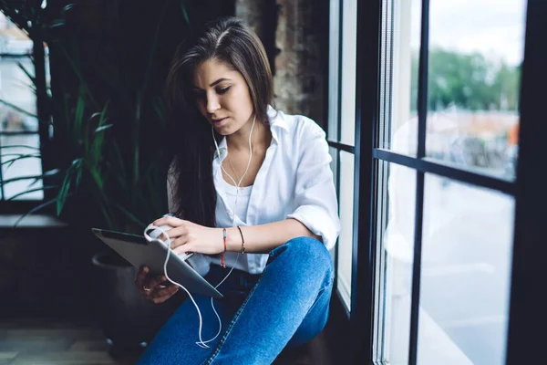 全神贯注的年轻女性 一头乌黑的长发 随意地穿着 带着耳机的平板电脑坐在窗台上 带着植物坐在创意的工作空间里 — 图库照片