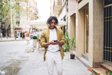 Handsome young male with curly hair using mobile phone on urban settings sending text messages and mails, positive 20s hipster guy browsing web page on smartphone searching destinations and location