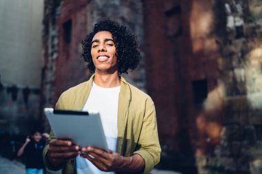 Low angle of positive confident young African American man in casual outfit smiling while communicating on tablet and enjoying sunny day against shabby brick building