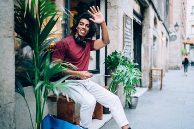 Side view cheerful young man in casual wear smiling and waving hand sitting on bench near green plants on street and looking away