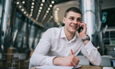 Cheerful man in formal shirt smiling and looking away while talking on mobile phone and taking notes during work in cafe