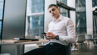 Young male freelancer in formal outfit surfing internet on cellphone while sitting at round table in modern cafe and looking at camera