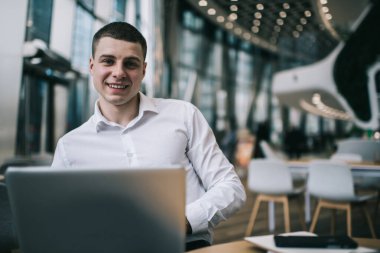 Cheerful male freelancer in formal clothes sitting in front of open netbook at table in modern cafeteria and looking at camera
