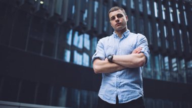 From below of serious young guy in casual clothes and wristwatch standing against modern building with crossed arms and looking at camera