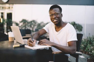Smiling African American freelancer making notes in diary while working on laptop in outdoor coffee shop on warm summer evening