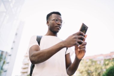 From below of busy pensive African American young male in white t shirt black backpack and eyeglasses attentively surfing smartphone while walking along city street