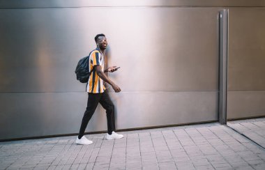 Side view of stylish hipster guy with travel backpack holding digital mobile phone and walking at urban street, millennial African American man in eyewear and earphones listening online music playlist