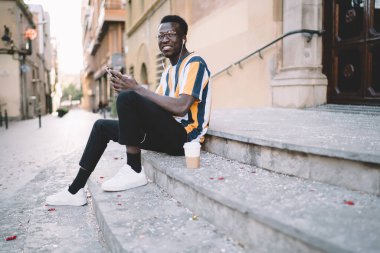 Portrait of millennial hipster guy using smartphone device during free time in city, dark skinned male blogger in earphones and classic eyewear smiling at camera holding cellphone technology