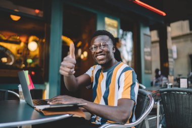 Portrait of cheerful male programmer in classic spectacles smiling at camera enjoying freelance lifestyle, happy African American hipster guy with thumbs up showing OK popularizes distance job
