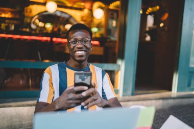 Portrait of funny male blogger with digital smartphone in hands sitting in street cafe and smiling at camera, toothy hipster guy in optical eyewear using mobile phone and cropped laptop computer